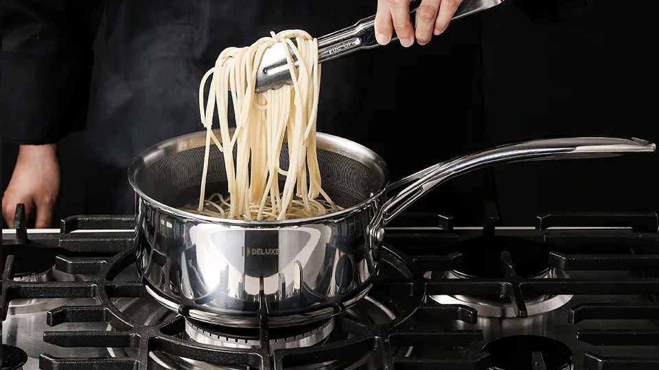 Person lifting long noodles with tongs from a shiny metal sauté pan on a stovetop, steam rising.
