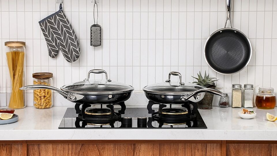 Two Deluxe Cookware black frying pans with glass lids on a modern induction stove top in a bright kitchen