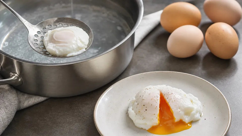 Slotted spoon lifts a poached egg from a Deluxe Cookware stainless pot; plated egg shows runny yolk beside brown eggs